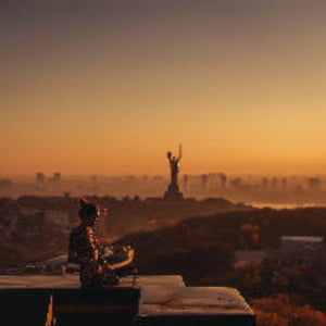 Person sitting on a ledge with a cityscape and statue in the background during sunset.